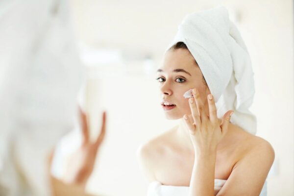 a fair-skinned brunette woman wearing a white towel uses a creamy lotion from her custom skincare routine